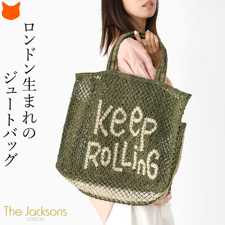 ロンドン生まれのジュートバッグ。前向きなメッセージ「Keep Rolling」がデザインされた、落ち着いたオリーブグリーン色の大容量で丈夫なザ・ジャクソンズの手編み大きめトートバッグを肩掛けする女性